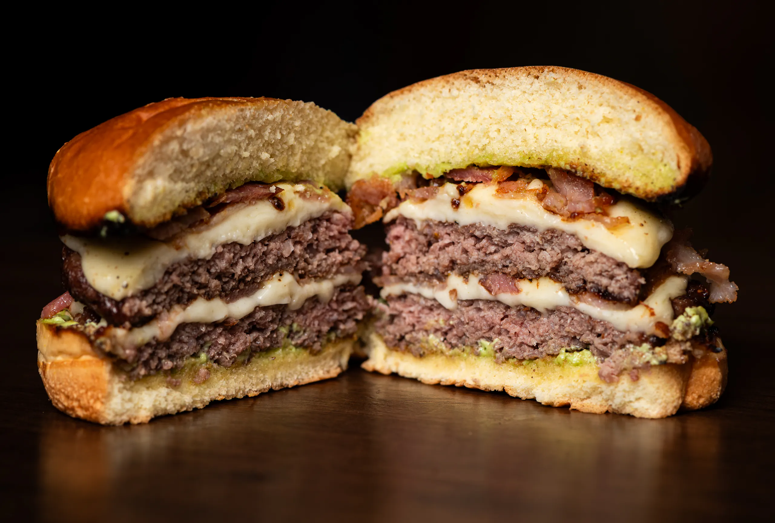 Brous Duplo Bacon cortado ao meio: duas carnes bovinas, queijo derretido entre elas, bacon e maionese verde sobre o pão selado.