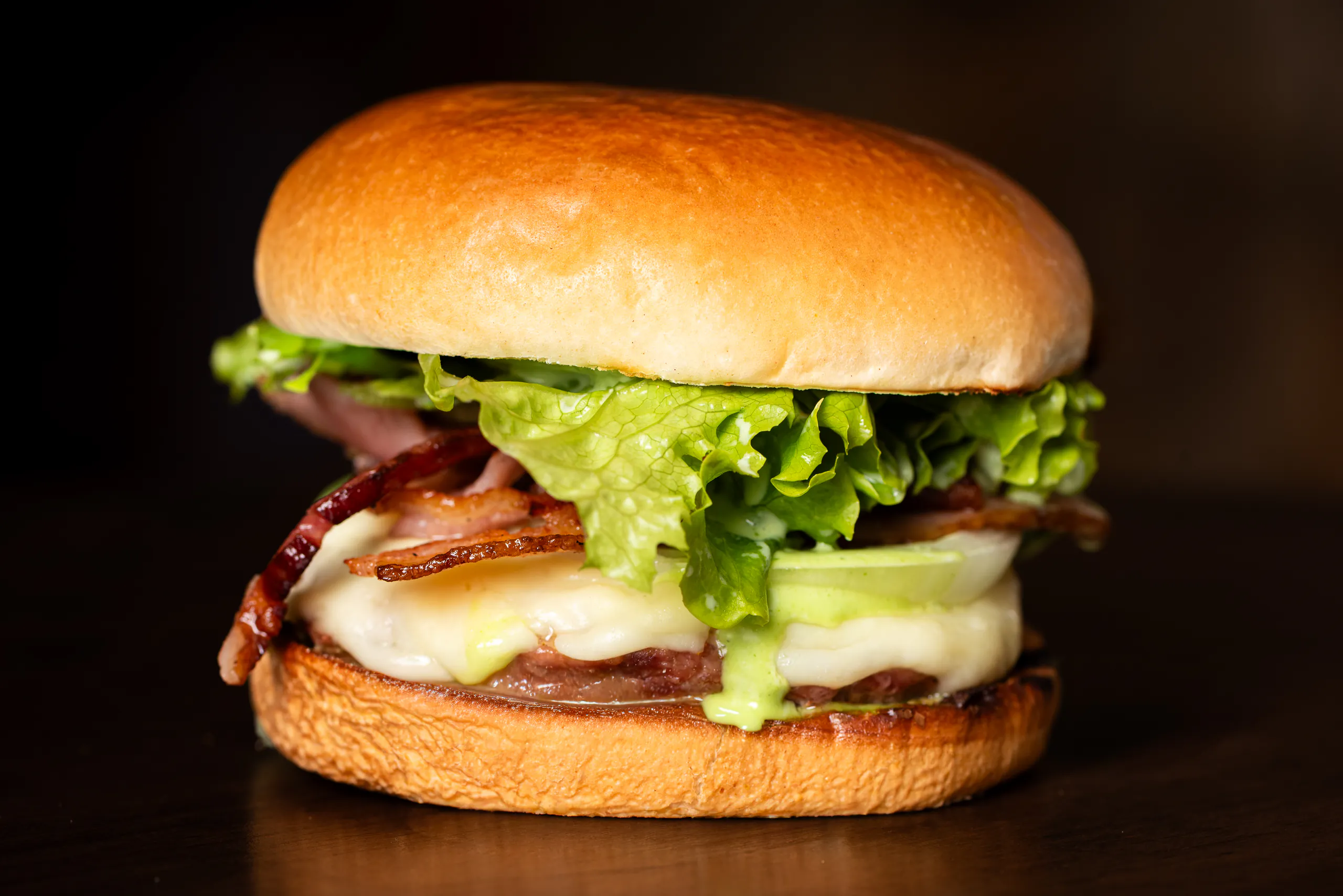 Pork Bacon Salada de frente, em close: pão selado, alface crespa, bacon, cebola, queijo derretido e maionese verde escorrendo sobre a carne suína.
